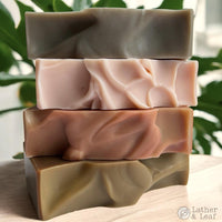 Stack of four different colored soap bars on a wooden surface with a blurred green leafy background.
