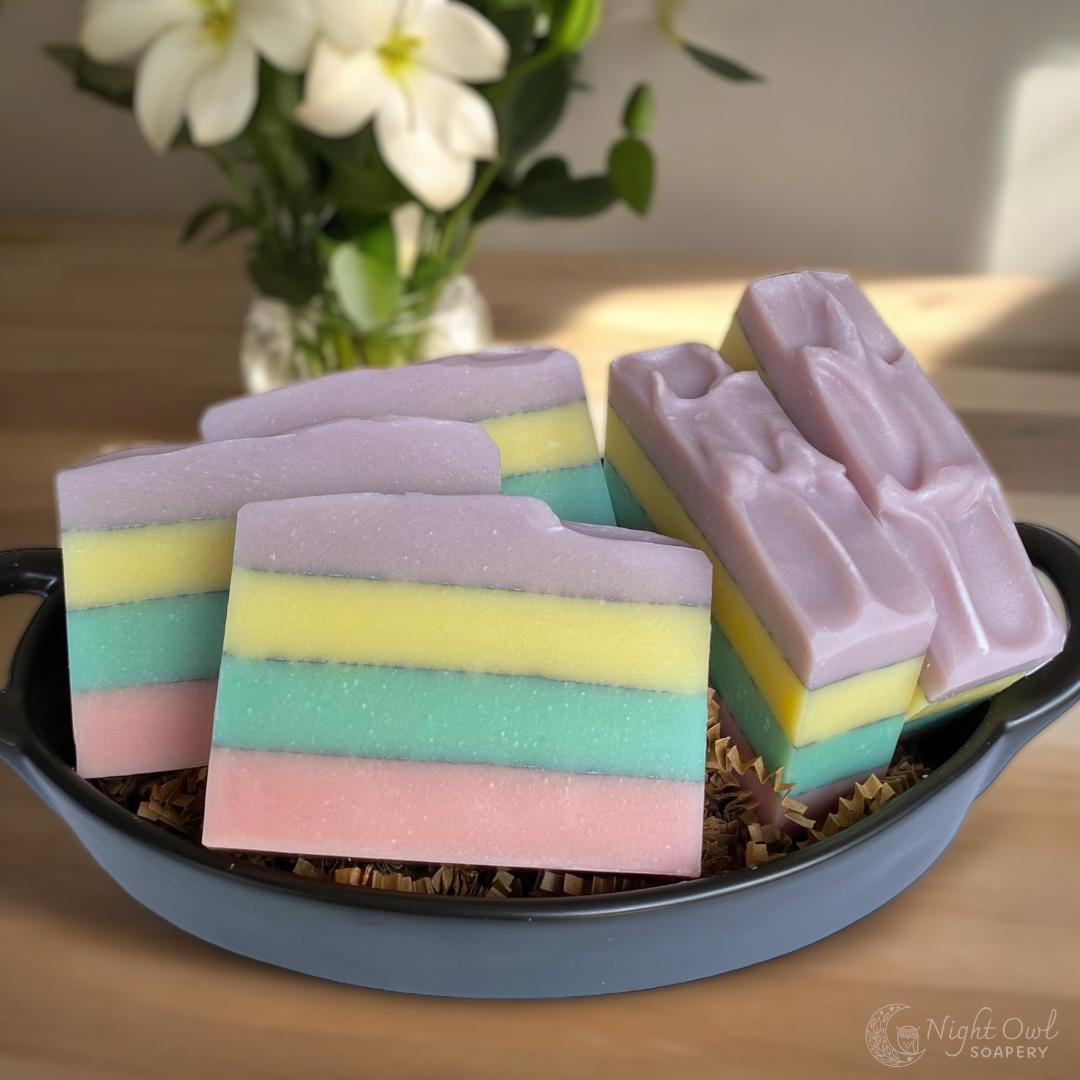 Colorful layered soap bars in a black dish with a vase of flowers in the background