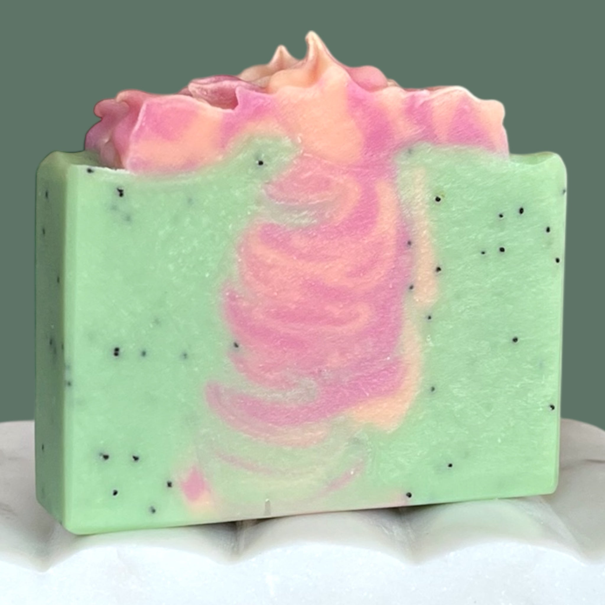 Light green bar of soap with pink and orange swirls and poppy seeds on a white surface