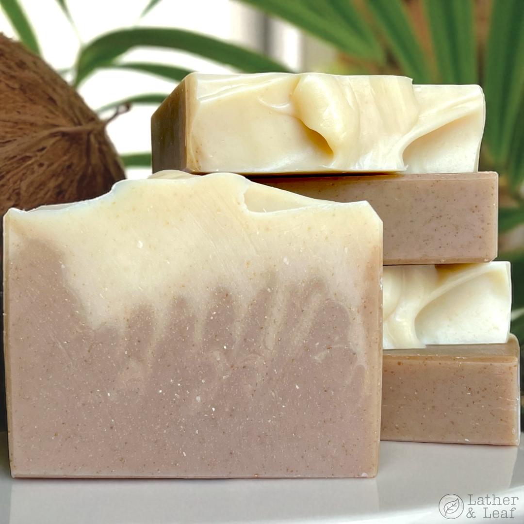 Three bars of soap stacked on a white surface with a coconut and green leaves in the background.