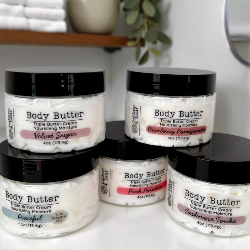 Collection of Lather & Leaf body butter jars on a white surface with a spa-like blurred background.