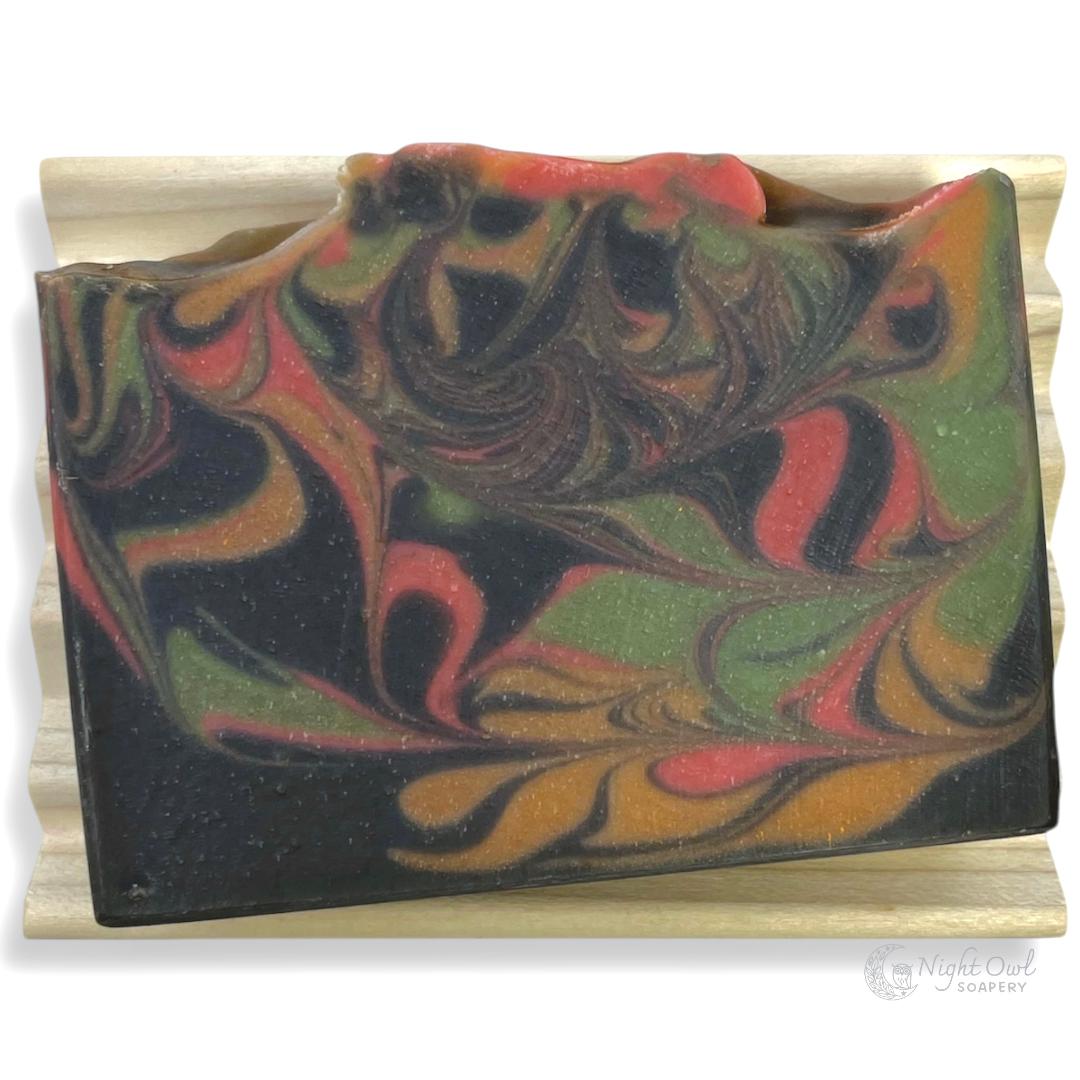 Black, gold, red and green soap bar with a swirled pattern on a wooden soap dish