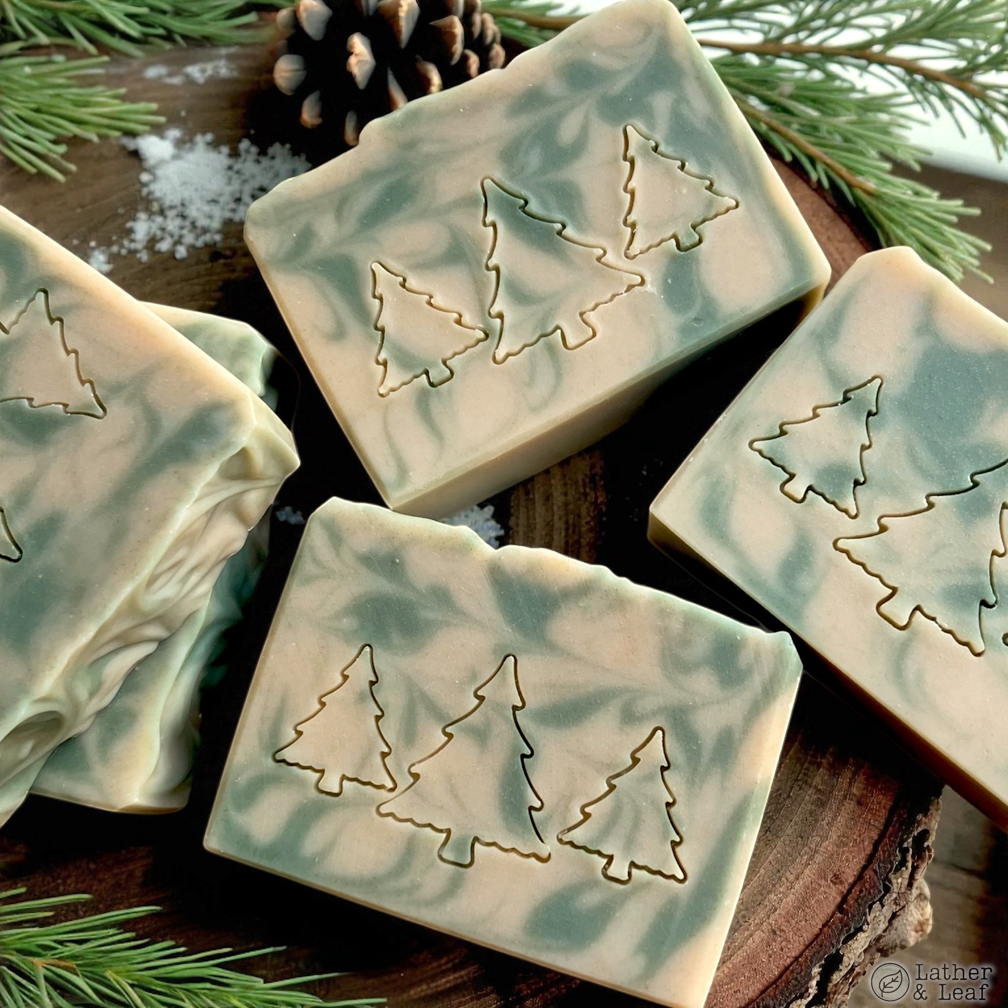 Handmade soap bars stamped with tree designs on a wooden surface with pine branches and snow.