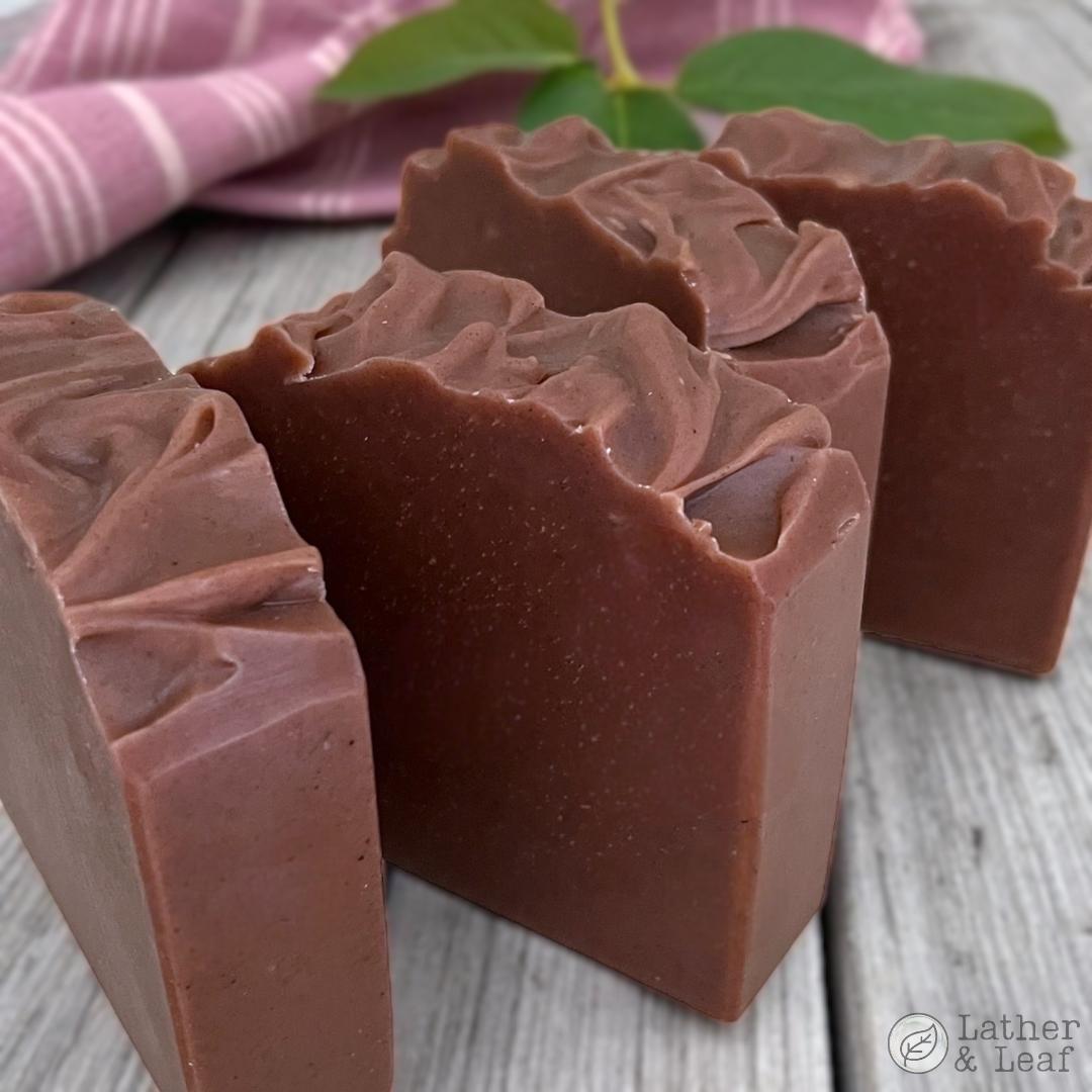 Three bars of maroon soap with a textured surface on a wooden surface, with a pink cloth and green leaves in the background.