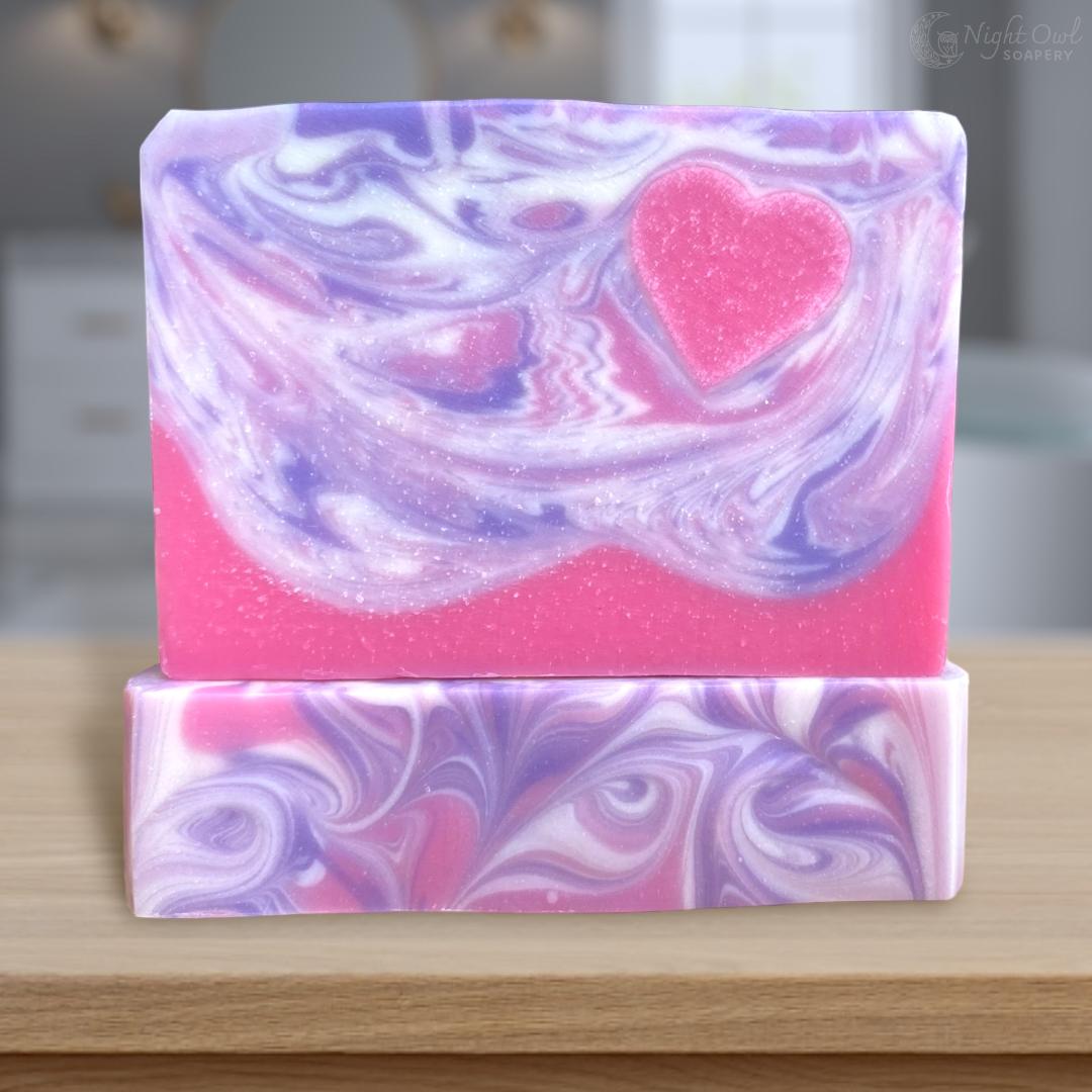 Two swirled soap bars with a pink heart embed design on a wooden surface