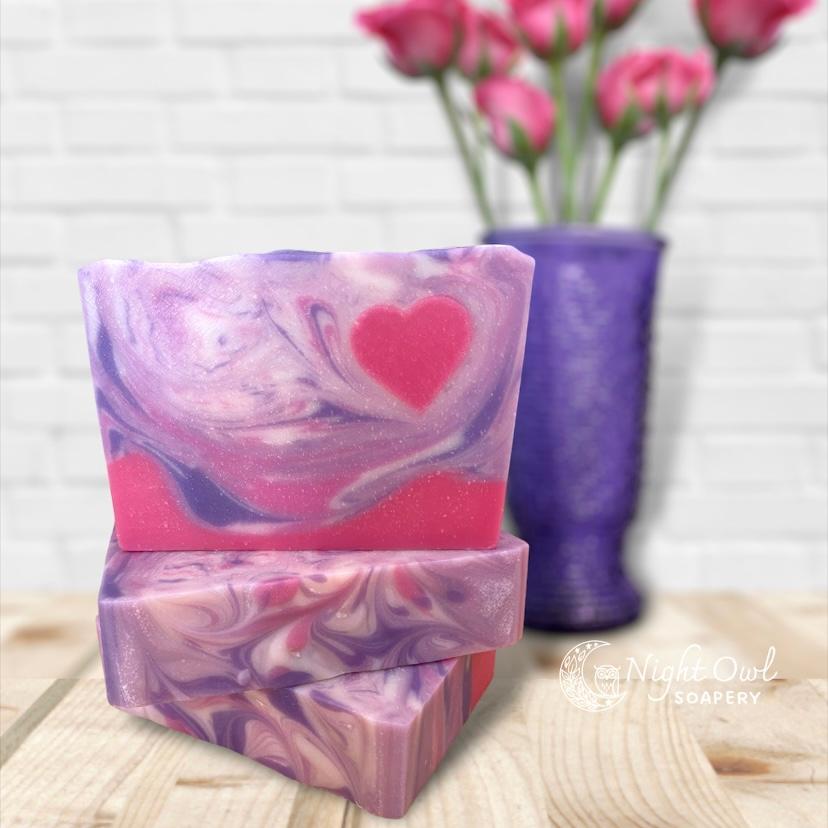 Stack of pink and purple swirled soap bars with a blurred vase of pink flowers in the background.