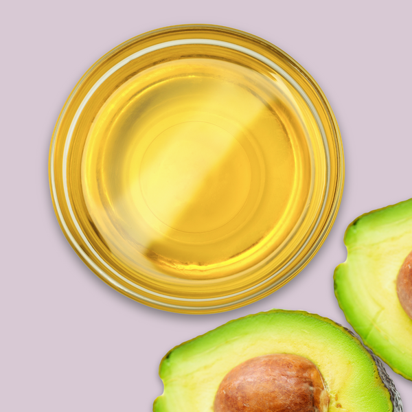 Glass bowl of avocado oil next to two halves of an avocado on a light purple background