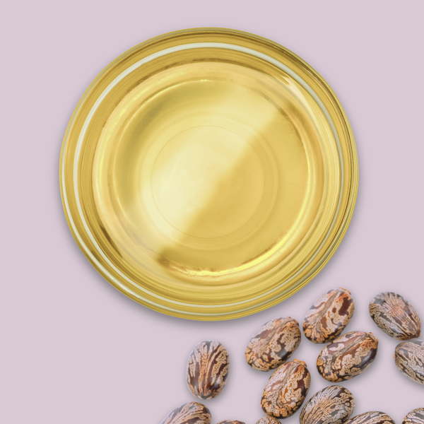 Glass bowl filled with castor oil on a light pink background with castor beans around it.