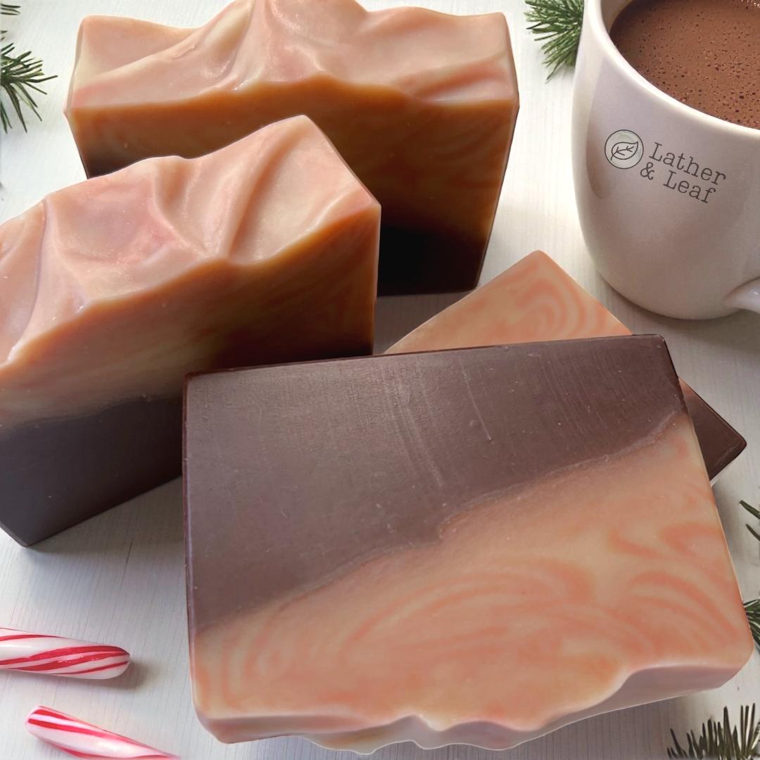 Layered soap bars with brown, red and white swirls, next to a mug of cocoa and candy cane pieces on a white surface.