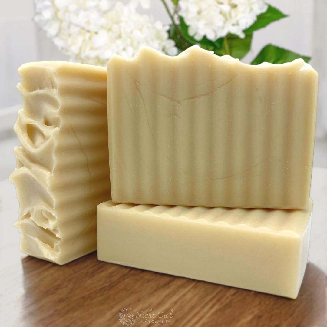 Three bars of soap stacked on a wooden surface with a blurred background of white lilacs
