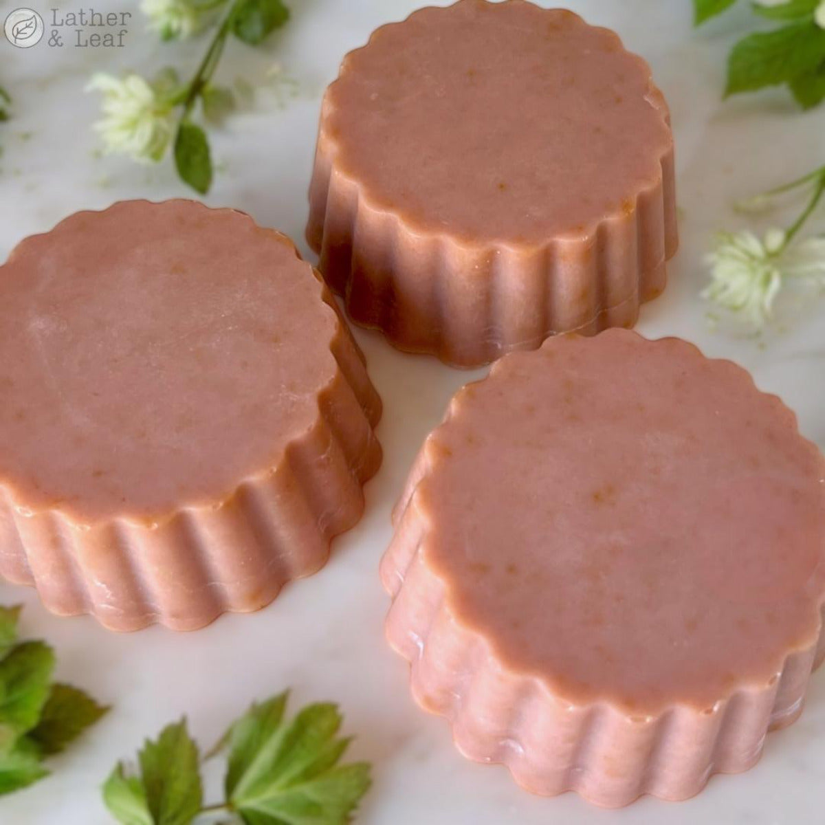 Three round bars of soap with a pinkish tint and ruffled edges, displayed on a surface with green leaves and white flowers around them.
