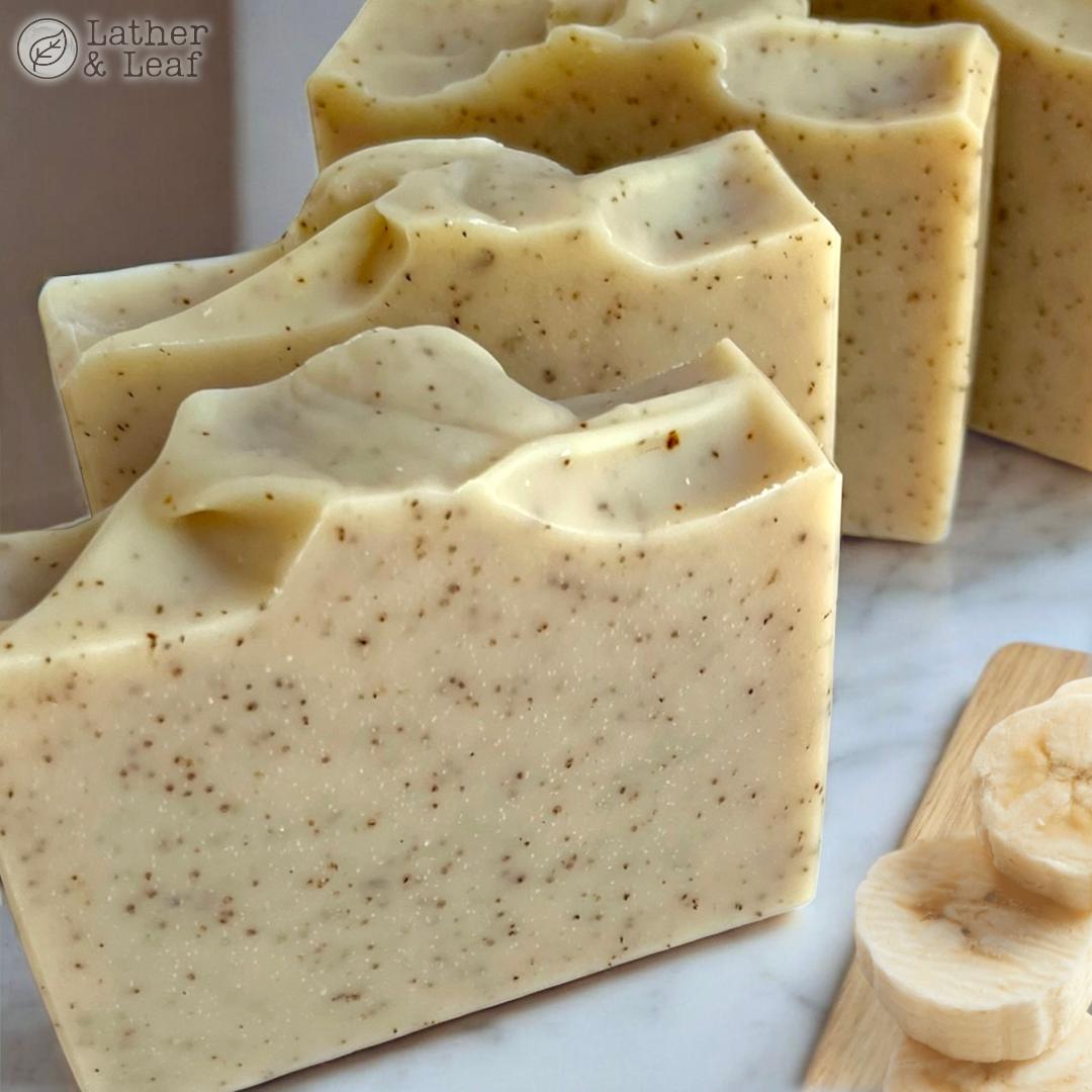 Close-up of beige soap bars with banana specks on a marble surface, next to sliced bananas on a wooden cutting board