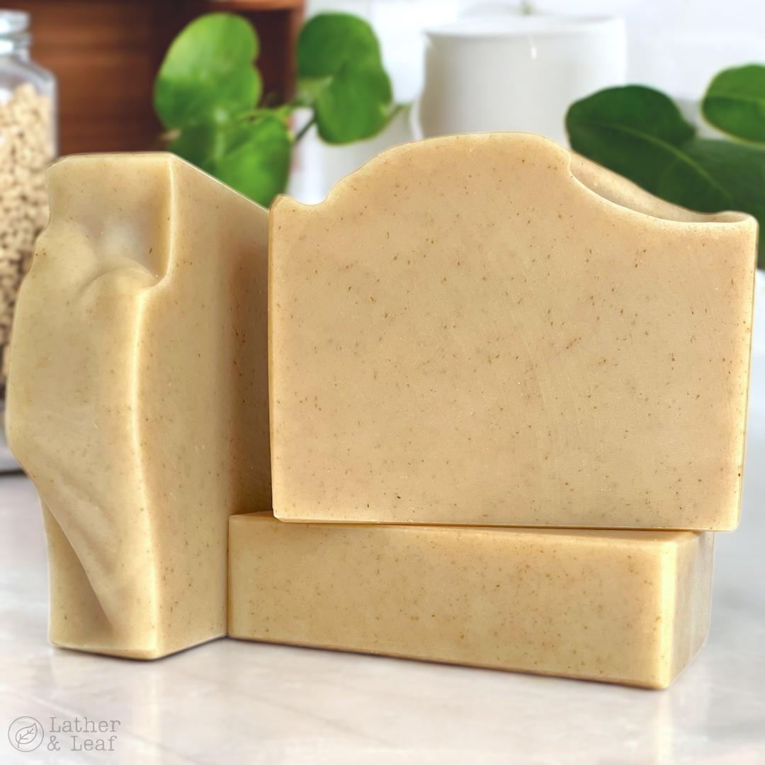Three bars of beige soap stacked on a light surface with blurred greenery in the background.