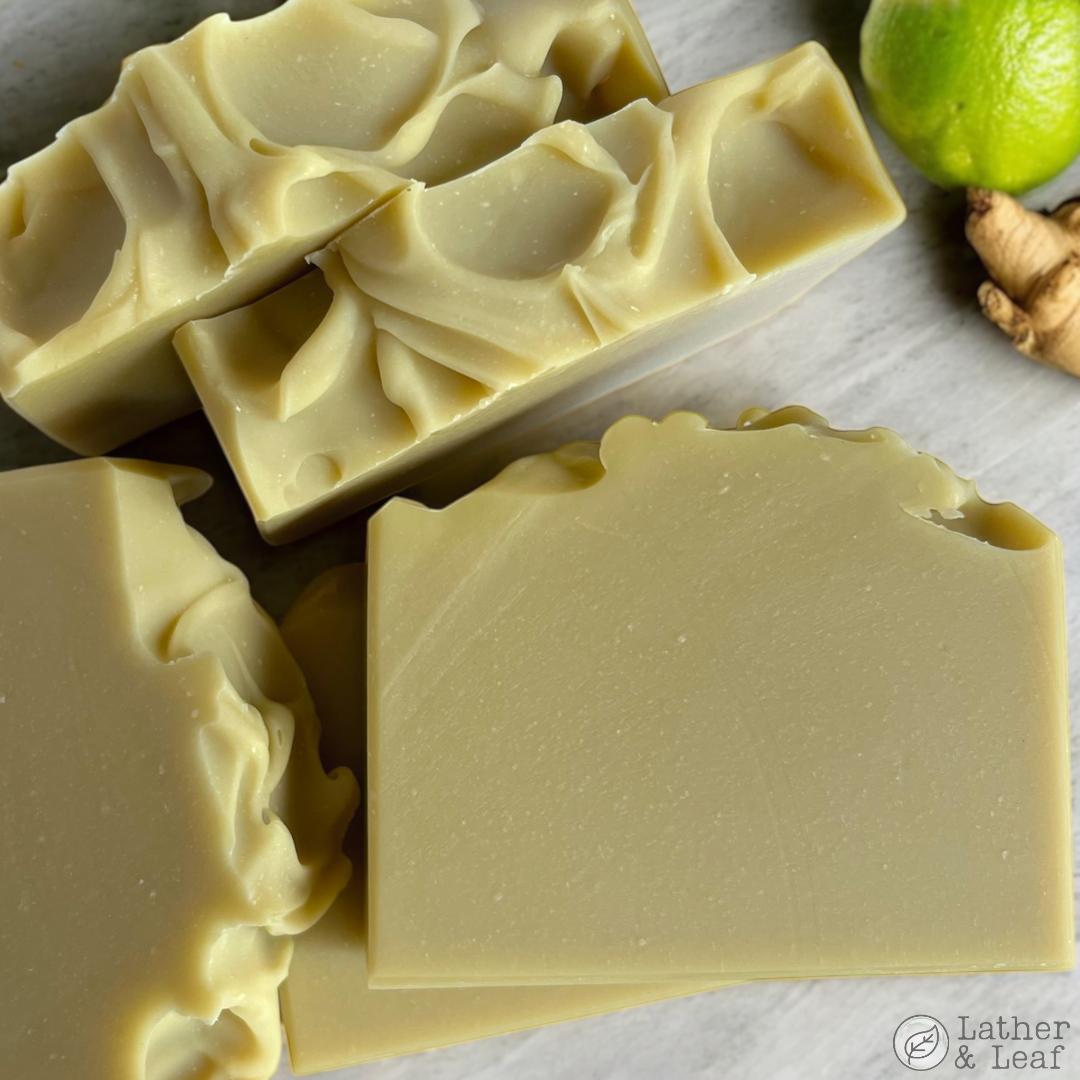 Bars of soap with decorative edges on a marble surface, with a lime and ginger root in the background.