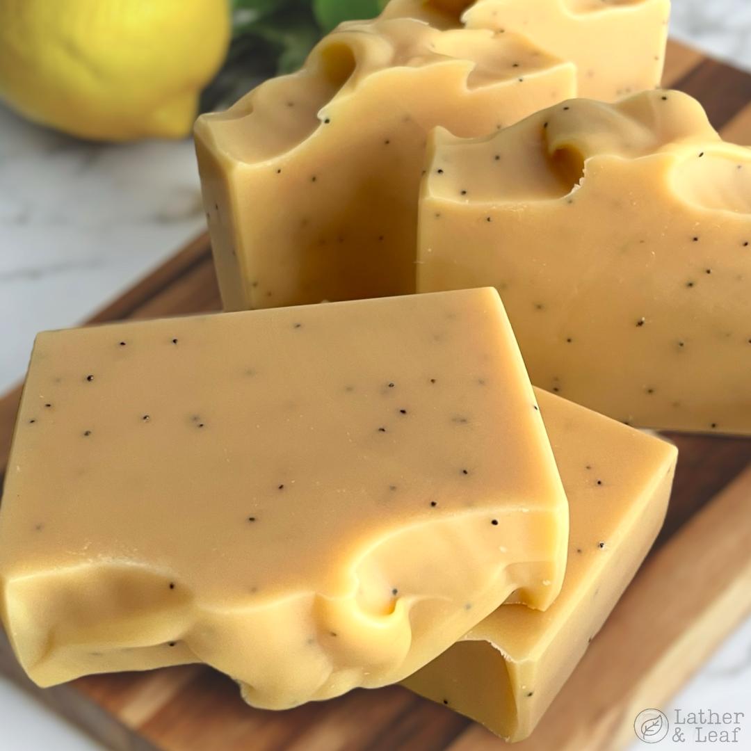 Bars of yellow soap with poppy seeds on a wooden board, with a lemon and leaves in the background.